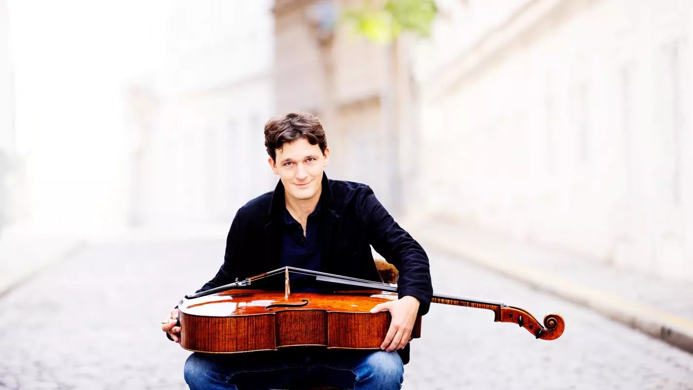 Man in black shirt and jeans sitting on a chair in the middle of a cobbled street holding a cello on his lap