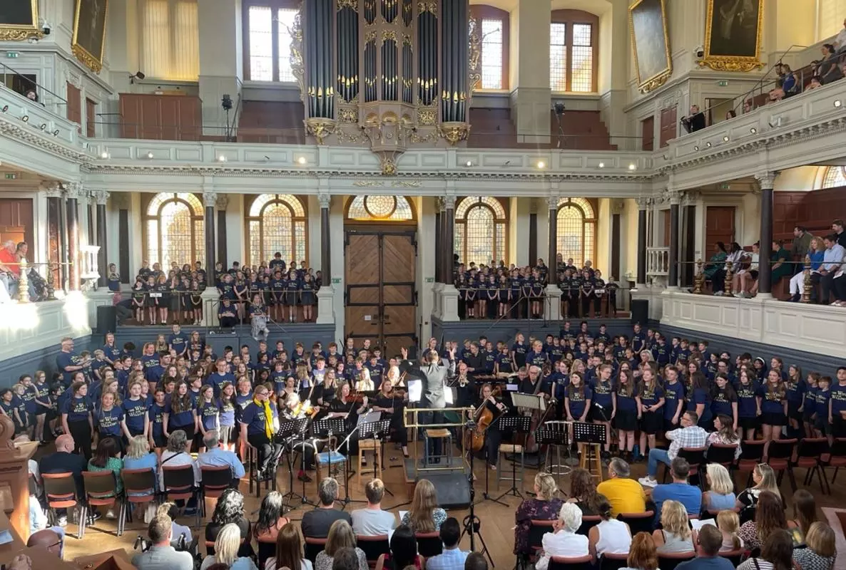 A choir and small orchestra congregate in the auditorium of the Sheldonian Theatre as an audience watches from both the floor and the galleries. 